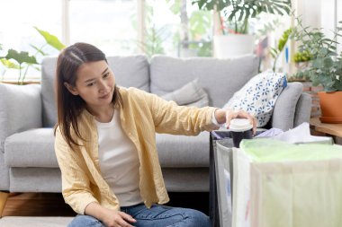 Asyalı genç bir kadın evde oturuyor kağıt ve plastik atıkları ayırıyor. Geri dönüşüm için çöp ayıklama. Küresel ısınmanın azaltılması. Çevre koruma kavramı.