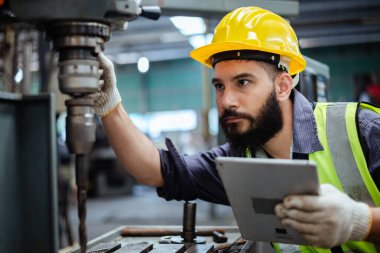 Teknisyen, metal fabrikasının elektrikli matkap mekanizmasını çalıştırmak için tablet kontrolü kullanıyor. Erkek işçi sarı kasketli, demir sondaj makinesi takıyor..