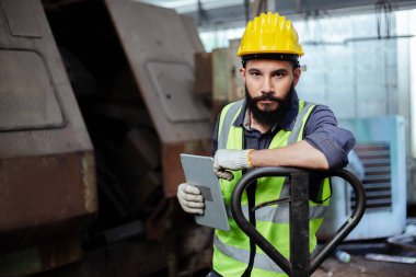 Mühendis portresi elinde tablet ve üniforma olan sarı kaskla torna fabrikasında duran kameraya bakıyor..