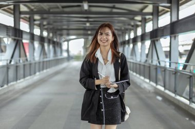 Siyah takım elbiseli çekici genç bir kadın elinde tabletle halka açık yerlerde kameraya bakıp gülümsüyor. Yaşam tarzı insan teknolojisi kavramı. Üniversitenin dışında bir kız öğrenci.