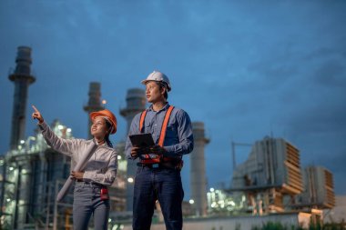 Asyalı ekip elemanı ve kadın mühendis gece yarısı petrolle çalışan elektrik santralinin önünde planlarla çalışıyorlar. İşçiler kule inşaatında çalışan üniformalı miğfer giyerler..