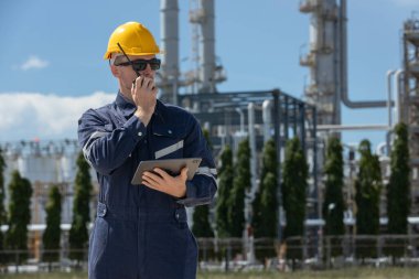 Profesyonel mühendis güvenlik üniforması ve rafine petrol petrokimya endüstrisinin yakınında bulunan tablet ve telsiz çalışma alanı tutan beyaz başlık giyer. Fabrika sitesi enerji santrali.