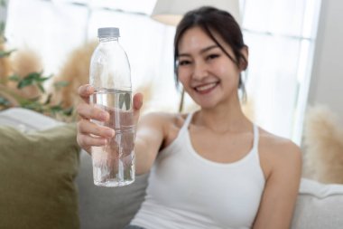 Su şişesini kapat. Oturma odasında egzersiz yaparken kanepede oturan Asyalı genç bayan sportif içme suyu. Her gün susuz kalma. Sağlıklı, sağlıklı, sağlıklı, sağlıklı bir kadın. Su içmek iyi hissettirir..