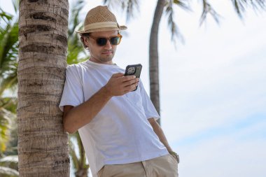 Güneş gözlüklü yakışıklı, beyaz şapkalı, akıllı telefon kullanan, tropik sahilde sohbet eden yaz tatilinde palmiye ağacının altında duran beyaz tişörtlü yakışıklı adam.. 