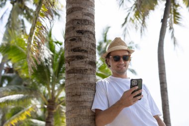 Güneş gözlüklü yakışıklı, beyaz şapkalı, akıllı telefon kullanan, tropik sahilde sohbet eden yaz tatilinde palmiye ağacının altında duran beyaz tişörtlü yakışıklı adam.. 