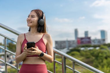 Genç ve güzel kadın spor kıyafetleri akıllı telefon kullanarak ve kulaklıkla müzik dinleyerek egzersiz yaparken mavi gökyüzü arka planında dinleniyor. fitness sağlıklı konsept.