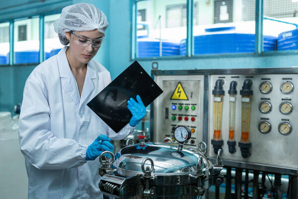 Female worker holding clipboard take note checking quality drinking water management system before process of filling water into bottles to bring out to consumers. Water production line of factory.