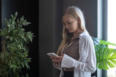 Güzel, beyaz tenli, sarı saçlı beyaz kadın ofisin penceresinde akıllı telefon kullanıyor..