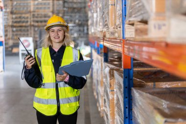 Kadın sarışın, profesyonel, güvenlik üniforması giyen, kasketli, pano tutan ve depodaki raflarda bulunan eşyaları inceleyen bir işçi. İşçi fabrikada hisse senedi denetimi yapıyor..