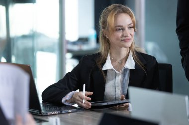 Genç profesyonel bir kadın konferans masasında elinde tabletle oturuyor ve bir iş toplantısına katılırken gülümsüyor. Doğal ışık modern ofis alanını doldurur..