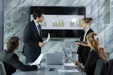 Bir grup profesyonel performans ölçümleri ve şık ofis ortamında gelecekteki hedefleri tartışıyor. Ekip, işbirliğini vurgulayarak ekranda gösterilen çizelgelerle meşgul oluyor.