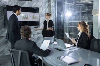 Takım üyeleri, çağdaş bir ofisteki şık bir konferans masasında otururken veri analizi ve performans ölçümleri sunan bir meslektaşı dinliyor..