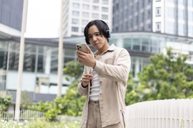 Şehir caddesinde yürüyen ve akıllı telefonlu müzik dinleyen genç adam kablosuz kulaklık takıyor. Mutlu erkek rahatlamak için sosyal ağ sohbetini kullanıyor..