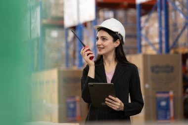 Kadın işçi, lojistikte çalışan stok mallarını kontrol etmek için telsiz sesli dijital tablet kullanıyor. Kadın profesyonel depo çalışanı güvenlik üniforması giyiyor ve depoda beyaz şapka denetleme ürünü var.. 