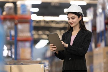 Kadın, dijital tablet kullanarak lojistikte çalışan hisse senetlerini kontrol ediyor. Kadın profesyonel depo çalışanı güvenlik üniforması giyiyor ve depoda beyaz şapka denetleme ürünü var.. 