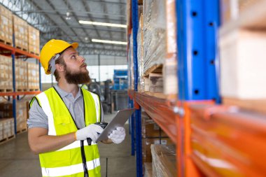 Depo işçisi elinde tablet tutarak depo deposundaki ürünlerin envanterini ve dağıtımını kontrol ediyor. Malların denetlenmesi ve yerleştirilmesi.