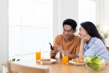 Çift birlikte gülerken yemek masasında portakal suyu ve kahvaltı malzemeleriyle akıllı telefonlara bakıyor..