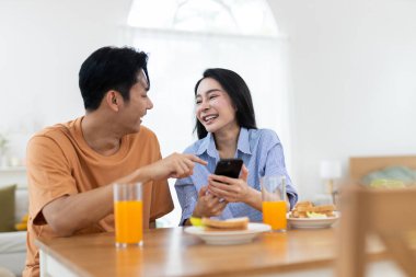 Çift birlikte gülerken yemek masasında portakal suyu ve kahvaltı malzemeleriyle akıllı telefonlara bakıyor..