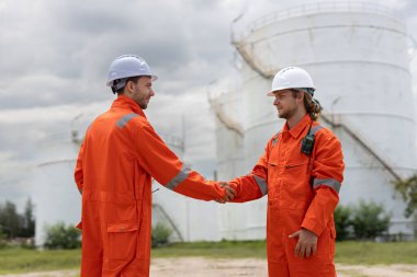 Turuncu takım elbiseli ve kasklı iki adam sanayi bölgesinde el sıkışıyor. Bulutlu gökyüzünün altında büyük bir depolama tankının yanında duruyorlar. Bu da takım çalışmasını ve işbirliğini gösteriyor..