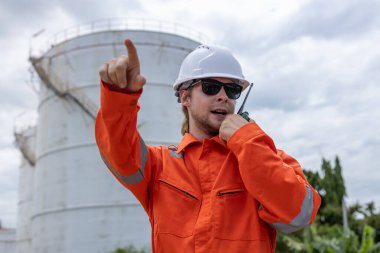 Turuncu tulumlu profesyonel mühendis ve telsiz telsizinden beyaz başlıklı adam ileri işaret ederken. Petrol petrokimya sanayii fabrikasında rafineride çalışıyorum..