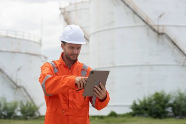 Petrokimya endüstrisi fabrikasının rafine edilmesi için endüstriyel depolama tanklarının yanında tablet kullanan güvenlik donanımı işçisi..