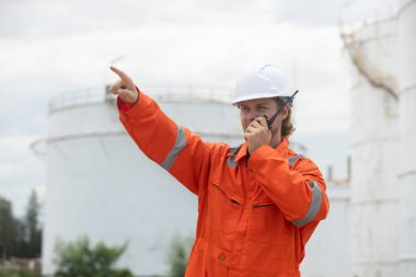 Turuncu zırhlı işçiler radyo aracılığıyla iletişim kuruyor, depolama tanklarıyla endüstriyel bir alanda faaliyetler yönetiyor..