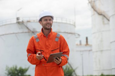 Petrokimya endüstrisi fabrikasının rafine edilmesi için endüstriyel depolama tanklarının yanında tablet kullanan güvenlik donanımı işçisi..