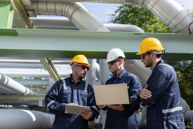Üç inşaat işçisi bir sanayi tesisindeki boru hatlarını denetlerken planları tartışıyor. Güvenlik kıyafeti giymişler ve gündüz vakti işlerine odaklanmışlar..