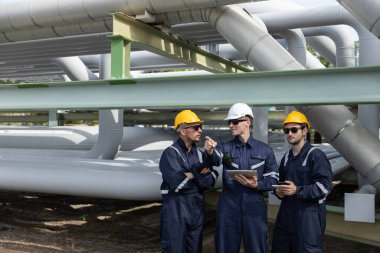 Üç inşaat işçisi bir sanayi tesisindeki boru hatlarını denetlerken planları tartışıyor. Güvenlik kıyafeti giymişler ve gündüz vakti işlerine odaklanmışlar..