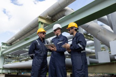 Üç inşaat işçisi bir sanayi tesisindeki boru hatlarını denetlerken planları tartışıyor. Güvenlik kıyafeti giymişler ve gündüz vakti işlerine odaklanmışlar..