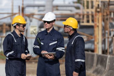 Güvenlik miğferleri ve koyu renk üniformalı üç işçi, sanayi bölgesinde tablet kontrol ekipmanları kullanarak bir tartışmaya giriştiler..