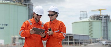 Turuncu takım elbiseli profesyonel mühendis ve beyaz başlıklı adam sıvılaştırılmış doğalgaz tesisi endüstriyel yapıları petrolünde tablet kullanıyor. Gaz nakil projeleri için araştırma planı