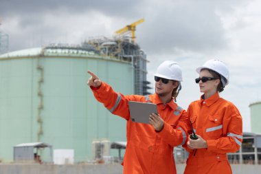 Turuncu takım elbiseli profesyonel mühendis ve beyaz başlıklı adam sıvılaştırılmış doğalgaz tesisi endüstriyel yapıları petrolünde tablet kullanıyor. Gaz enerji nakil projeleri için araştırma planlama