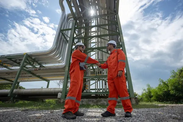 İnşaat alanında turuncu güvenlik donanımı giyen iki mühendis, büyük boru hatlarıyla el sıkışıyor. Gün ışığını aydınlatıyor, takım çalışması ve işbirliği sergiliyorlar..