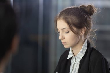 Açık kahverengi saçlı bir kadın, modern ofis ortamındaki bir toplantı sırasında dikkatle dinliyor. Masanın karşısından bir erkek arkadaşı onunla konuşuyor..