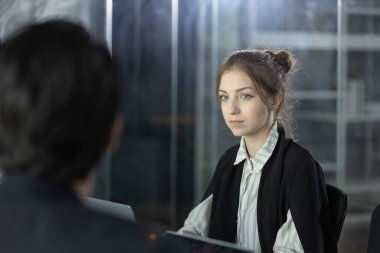 Açık kahverengi saçlı bir kadın, modern ofis ortamındaki bir toplantı sırasında dikkatle dinliyor. Masanın karşısından bir erkek arkadaşı onunla konuşuyor..