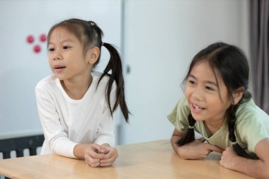 İki genç kız kardeş iyi aydınlatılmış bir odada ahşap bir masada dikkatle konuşuyorlar. Pencerelerden gelen doğal ışık sıcak atmosferi güçlendiriyor ve davetkar bir alan yaratıyor..