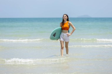 Neşeli genç kadınlar ellerinde sörf tahtasıyla güneşli sahilde sudan çıkıyorlar. Parlak turuncu bluz ve beyaz şort giyiyor. Arka planda açık mavi gökyüzü var..