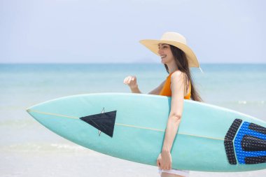 Neşeli genç bir kadın elinde sörf tahtasıyla güneşli sahilde sudan çıkıyor. Parlak turuncu bluz ve beyaz şort giyiyor. Arka planda açık mavi gökyüzü var..