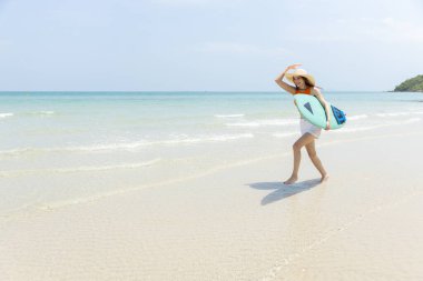 Neşeli genç bir kadın elinde sörf tahtasıyla güneşli sahilde sudan çıkıyor. Parlak turuncu bluz ve beyaz şort giyiyor. Arka planda açık mavi gökyüzü var..