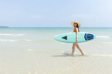 Neşeli genç bir kadın elinde sörf tahtasıyla güneşli sahilde sudan çıkıyor. Parlak turuncu bluz ve beyaz şort giyiyor. Arka planda açık mavi gökyüzü var..