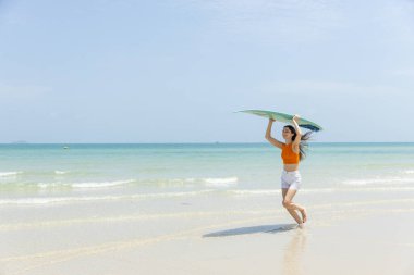 Neşeli genç bir kadın elinde sörf tahtasıyla güneşli sahilde sudan çıkıyor. Parlak turuncu bluz ve beyaz şort giyiyor. Arka planda açık mavi gökyüzü var..