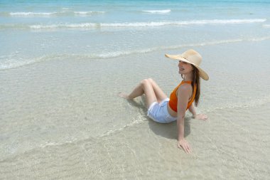 Hasır şapkalı, turuncu şortlu, beyaz şortlu, mavi gökyüzü ve turkuaz deniz manzaralı tropikal bir yerde oturan genç kadınlar..