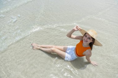 Hasır şapkalı, turuncu şortlu, beyaz şortlu, mavi gökyüzü ve turkuaz deniz manzaralı tropikal bir yerde oturan genç kadınlar..