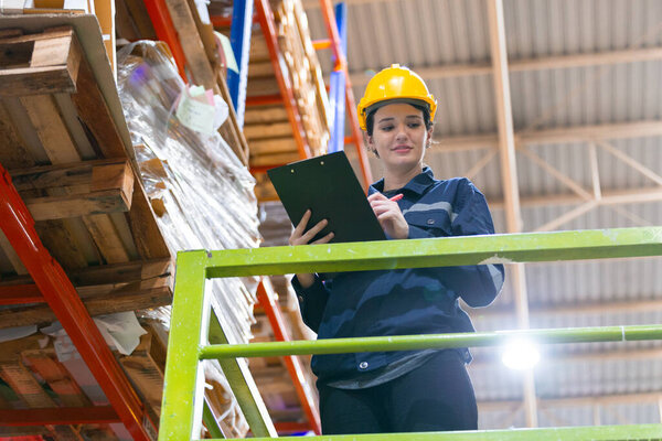 Women warehouse worker checking product cardboard on shelf in storage. Large industry in paper product line manufacturing. Employees wear uniform and helmet yellow counting stock on shelves.