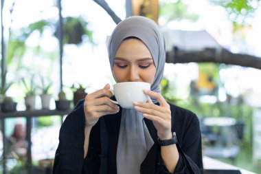 Başörtüsü takan gülümseyen kadın elinde bitki ve doğal ışıkla dolu bir kafede oturan bir fincan kahve tutuyor. Atmosfer sıcak ve davetkar..
