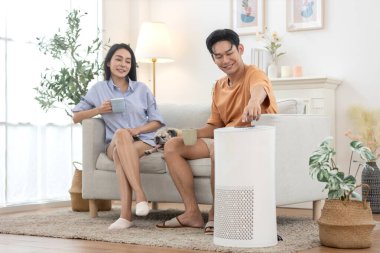 Genç Asyalı çift sabah kahvesini içip kanepede rahatlıyor oturma odasında, yanında küçük köpekleri dinleniyor, adam evin temiz havası için güç düğmesine basıyor..