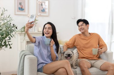 Mutlu genç Asyalı çift sabah kahvesini içip kanepede rahatlıyor. Oturma odasında gülümsüyor. Akıllı telefon kullanıyor..