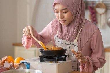 Mutfakta tavada yemek pişirmekten mutlu olan tesettürlü modern Müslüman kadın, sağlıklı helal tarzda lezzetli tatların tadını çıkarıyor, İslami yaşam özgürlüğü.