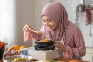 Modern mutfakta tesettürlü, mutlu yemek yapan, tencereye tahta kaşıkla yemek yiyen, taze sebzeler ve meyveler yerleştiren Müslüman bir kadın. Kepçelemek ve yemeği tatmak keyifli..
